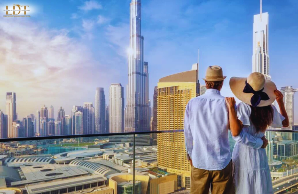 A couple in summer attire stands on a balcony overlooking the modern skyscrapers and cityscape of Dubai, including the Burj Khalifa, contemplating if Dubai is costly for Indians on a sunny day.