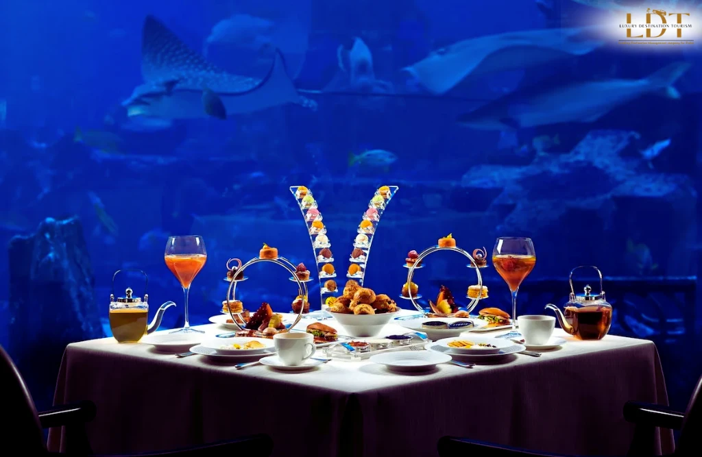 An elegant, fully set dining table with pastries, tea, and desserts is placed before a vast aquarium window teeming with fish—including a whale shark—offering an incredible underwater dining experience. Is food in Dubai expensive? This setting says it’s worth every dirham.